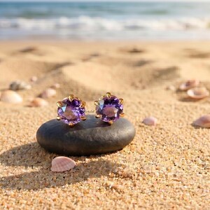 May include: A pair of round-cut purple amethyst stud earrings set in gold prongs. The earrings are resting on a smooth, dark gray oval stone. The faceted purple gemstones catch the light, displaying deep violet hues and clear, reflective surfaces.