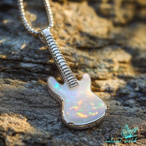 Puede incluir: Un colgante de guitarra plateada con un cuerpo de ópalo iridiscente cuelga de una delicada cadena. El cuerpo del colgante tiene una base blanca nacarada con destellos azules, verdes y amarillos. El mástil y la pala de la guitarra son plateados.
