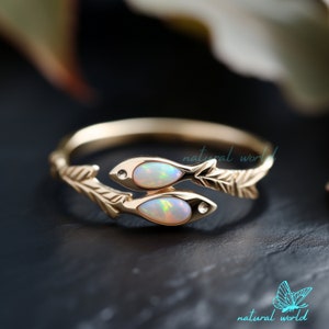 Può includere: Anello in oro con due disegni di pesci, ognuno con una pietra preziosa di opale. I pesci sono intrecciati con rami frondosi.