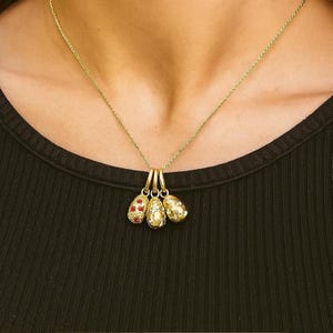 Puede incluir: Un collar dorado con tres colgantes. Los colgantes tienen forma de huevo, uno adornado con detalles rojos y los otros con un acabado dorado texturizado. El collar se lleva sobre una camiseta negra.