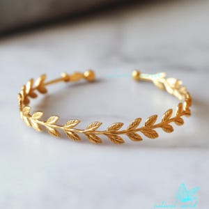 Pode incluir: Um bracelete dourado com um delicado design de folhas. O bracelete é aberto e ajustável.