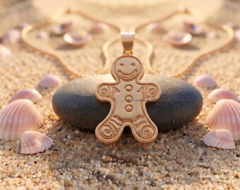 May include: A gold-toned gingerbread man pendant featuring a smiling face, three circular buttons on the torso, and swirl patterns on the arms and legs. The pendant is attached to a delicate gold chain and rests on a smooth, dark grey stone.