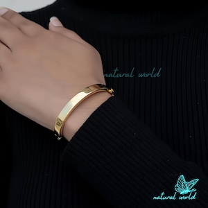 May include: A gold bangle bracelet with a hinged clasp. The bracelet is on a person's wrist.