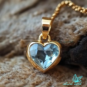May include: A gold heart-shaped pendant with a light blue gemstone. The pendant is on a gold chain.