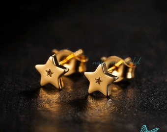 Puede incluir: Un par de pendientes de botón dorados con forma de estrella. Cada pendiente presenta un pequeño recorte de estrella en el centro. Los pendientes se muestran sobre una superficie oscura, con el texto "natural world" visible en el fondo.