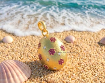 Pode incluir: Um pingente dourado em forma de ovo com acabamento fosco texturizado, apresentando padrões florais incrustados feitos de pequenas pedras preciosas vermelhas e pedras brancas transparentes, com uma argola circular no topo para corrente.