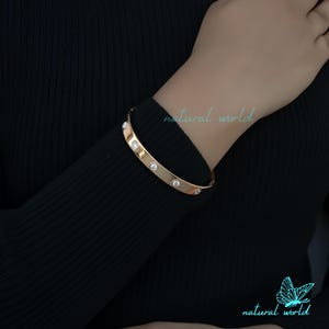 Könnte beinhalten: Ein goldenes Armreif-Armband mit klaren Strasssteinen, die in das Metall eingefasst sind. Das Armband ist offen und hat ein schlichtes Design.