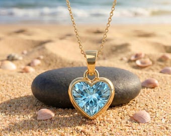 Puede incluir: Un collar de oro con un colgante en forma de corazón que presenta una piedra preciosa azul claro facetada. El colgante cuelga de una delicada cadena de oro, mostrando un engaste de metal pulido que enmarca la piedra azul central en corazón.