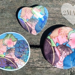 Pode incluir: Três botões coloridos com uma ilustração de três figuras se abraçando. Um botão tem formato de coração e os outros são redondos. As figuras têm cabelo loiro e verde e usam roupas azuis e roxas.