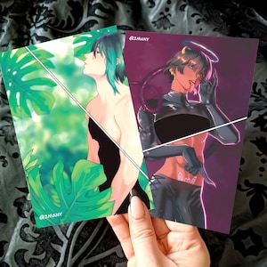 Op de afbeelding: Twee kunstdrukken met illustraties van figuren. Eén print toont een persoon met turquoise haar in een tropische omgeving, de andere toont een persoon met donker haar en een halo. De naam van de kunstenaar, @ZMANY, is zichtbaar op beide prints.