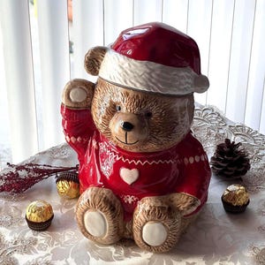 Puede incluir: Un tarro de galletas de cerámica con forma de oso de peluche que lleva un suéter rojo y un gorro de Papá Noel. El oso tiene un corazón blanco en su suéter y está rodeado de bombones dorados. Un adorno festivo.