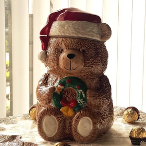 May include: A brown ceramic teddy bear cookie jar wearing a Santa hat and holding a Christmas wreath. The jar has a smiling face and is sitting on a table with gold-wrapped chocolates.