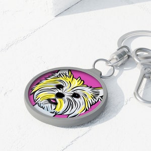 Op de afbeelding: Een ronde sleutelhanger met een illustratie van een Yorkshire Terrier in pop-art stijl. Het gezicht van de hond staat tegen een roze achtergrond, met een grijze rand. De sleutelhanger heeft een zilverkleurige ring en sluiting.