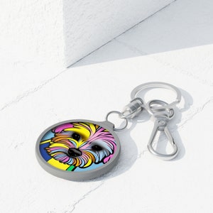 May include: A circular, grey keyring with a vibrant pop art style image of a dog's face. The dog's fur is rendered in yellow, pink, blue, and green hues. The keyring has a silver-toned ring and a clasp for attachment.