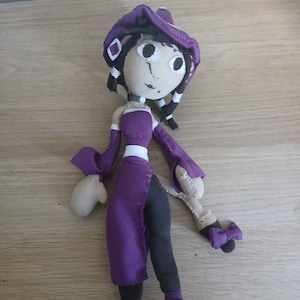 Könnte beinhalten: Eine handgefertigte Stoffpuppe mit einem violetten Hut, einem violett-schwarzen Kleid mit weißem Taillenband und dunklem Haar. Die Puppe hat große weiße Augen mit schwarzen Pupillen, einen gestickten Mund und trägt violett-schwarze Schuhe.