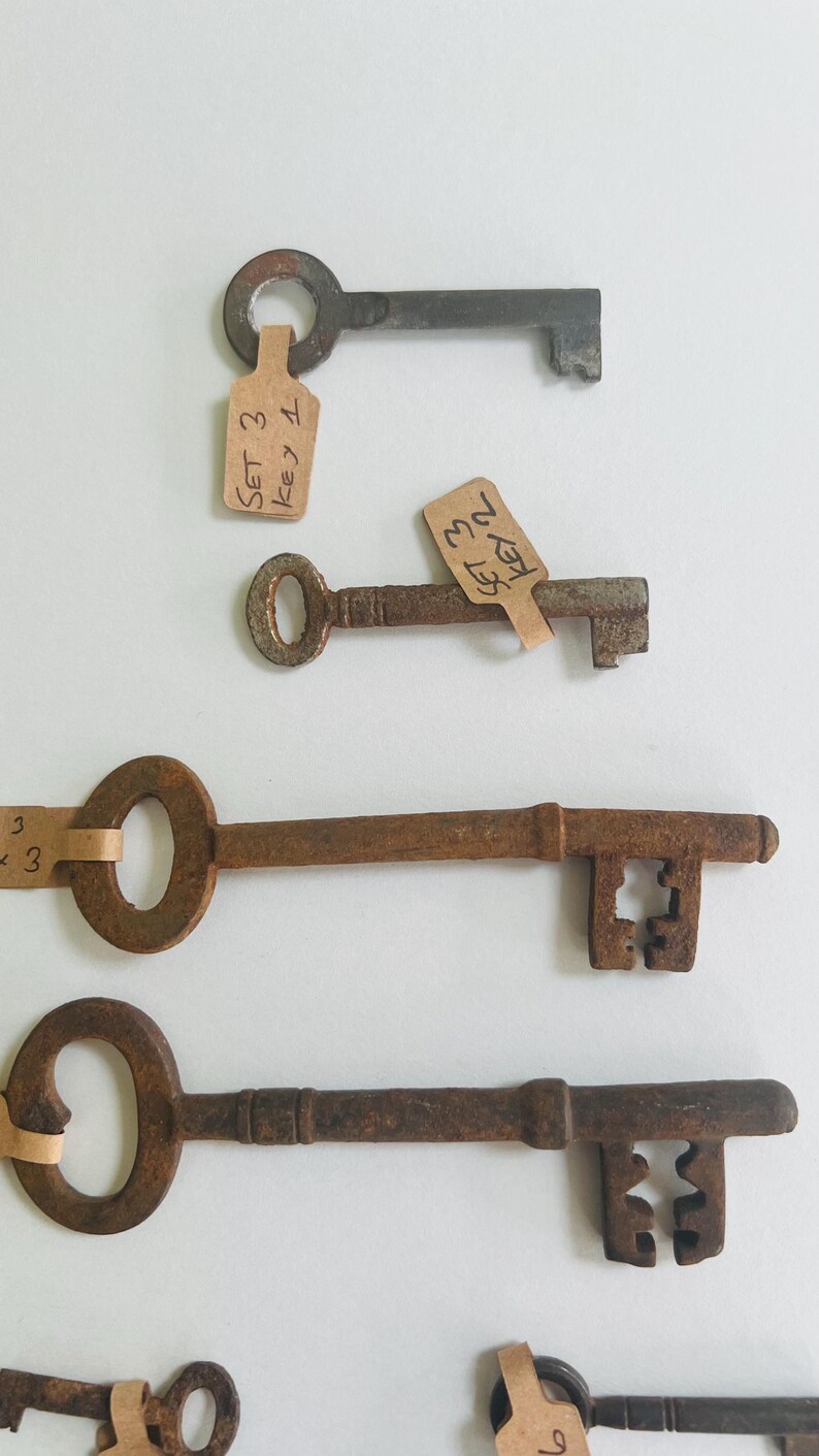 May include: Several antique keys with a rusty brown finish are arranged against a white background. Each key has a unique design, with some featuring decorative cutouts. Small tags with handwritten text are attached to some of the keys.