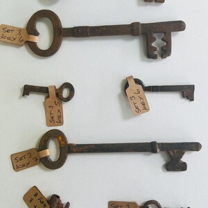 May include: Collection of antique metal keys with various designs and sizes. Each key is a rusty brown color and has a tag attached with handwritten text. The keys are arranged against a white background.