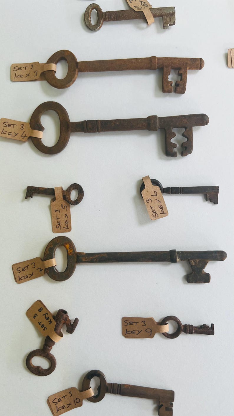 May include: Collection of antique metal keys with various designs and sizes. Each key is a rusty brown color and has a tag attached with handwritten text. The keys are arranged against a white background.