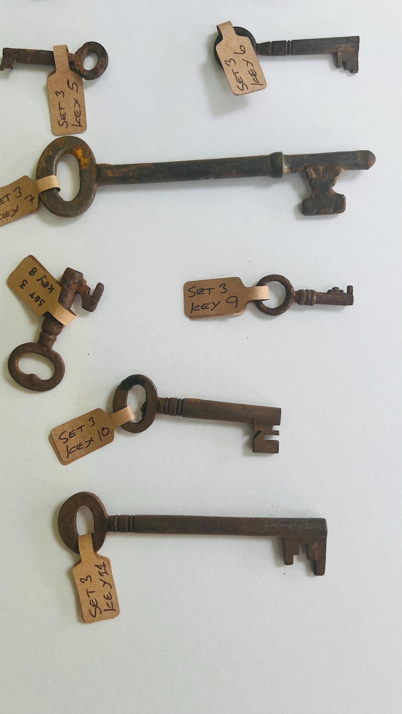 May include: Collection of antique keys with various designs and sizes. Each key is a rusty brown color and has a tag attached with handwritten text. The keys vary in shape, with some having ornate handles and intricate key bits.