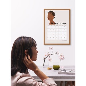 May include: A framed calendar with a brown frame and a beige background. The calendar features a stylized illustration of a woman with brown skin and dark hair. The month of November is displayed with the days of the week labeled.