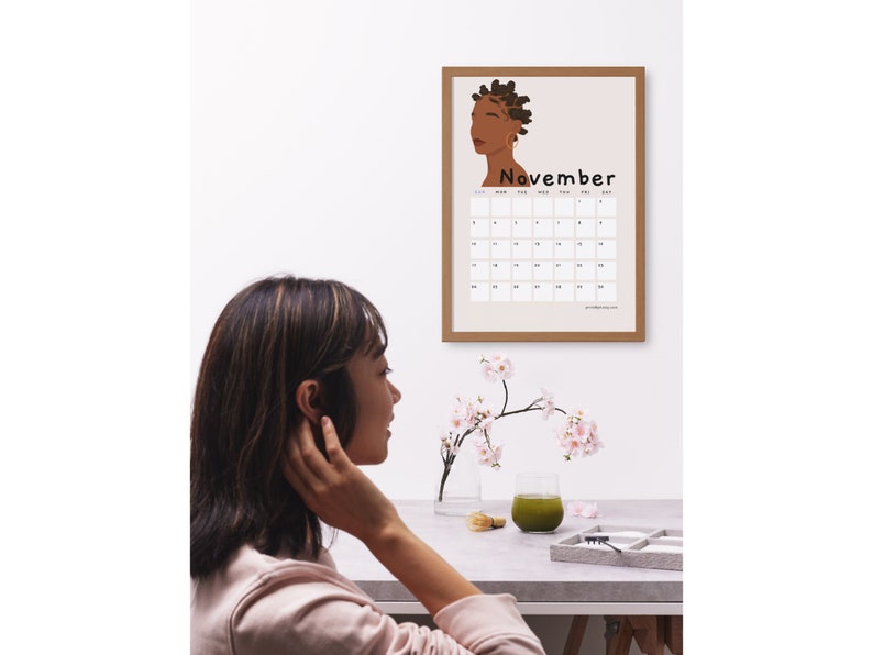 May include: A framed calendar with a brown frame and a beige background. The calendar features a stylized illustration of a woman with brown skin and dark hair. The month of November is displayed with the days of the week labeled.