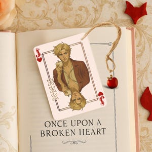 May include: A bookmark featuring a playing card design with an illustration of a person holding an apple. The card has a brown border, the Jack of Hearts symbol, and the text "ONCE UPON A BROKEN HEART". A red apple charm hangs from a twine string.