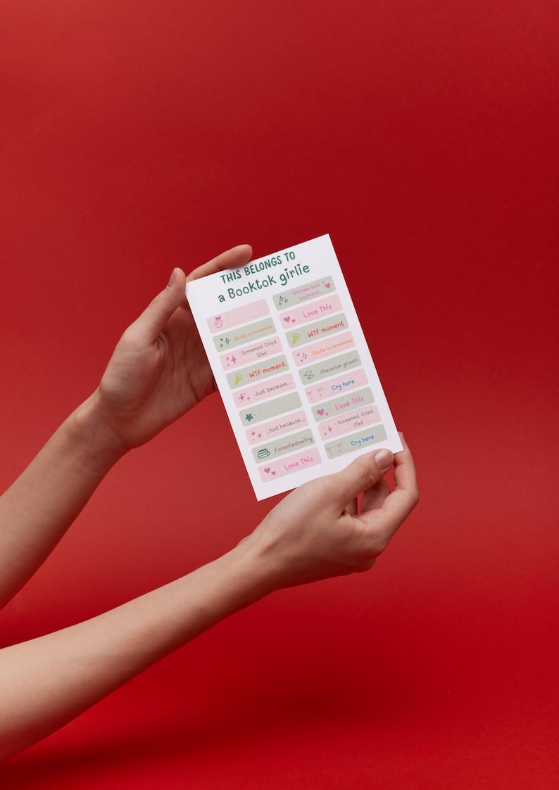 Op de afbeelding: Een witte kaart, vastgehouden door handen tegen een rode achtergrond. De kaart leest "THIS BELONGS TO a Booktok girlie" en bevat labels met zinnen als "WTF moment" en "Love This" in roze, groen en grijs.