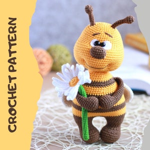 Può includere: Schema all'uncinetto per un simpatico giocattolo amigurumi a forma di ape che tiene un margherita bianca. L'ape è gialla e marrone con strisce nere e ha grandi occhi neri. Lo schema è disponibile per il download in PDF.
