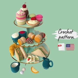 Puede incluir: Un expositor de postres de ganchillo de tres niveles con varias golosinas de ganchillo, que incluyen cupcakes, macarons, sándwiches y bolsitas de té. El expositor es azul claro y el fondo es verde. El texto "Crochet pattern" es visible.