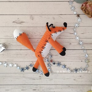 May include: A crocheted orange and white fox toy with black eyes and a stitched-on smile. The fox is lying on a light wood surface with a silver star garland.