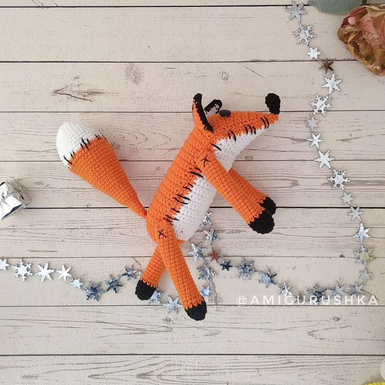 May include: A crocheted orange and white fox toy with black eyes and a stitched-on smile. The fox is lying on a light wood surface with a silver star garland.