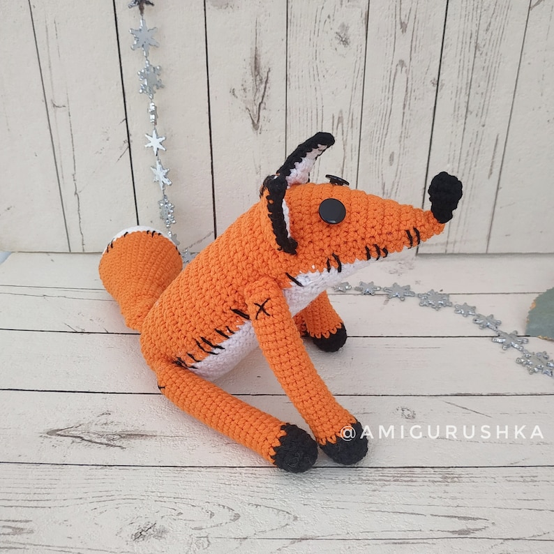 May include: A crocheted orange fox with black eyes and a white stitched belly. The fox is sitting on a white wooden surface.
