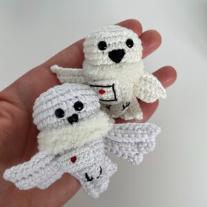 May include: Two handmade white crochet owl ornaments with black eyes and beaks are held in a hand. The owls have small wings and a fluffy white chest. One owl has a small red heart on its chest.
