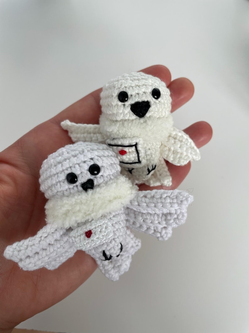 May include: Two handmade white crochet owl ornaments with black eyes and beaks are held in a hand. The owls have small wings and a fluffy white chest. One owl has a small red heart on its chest.