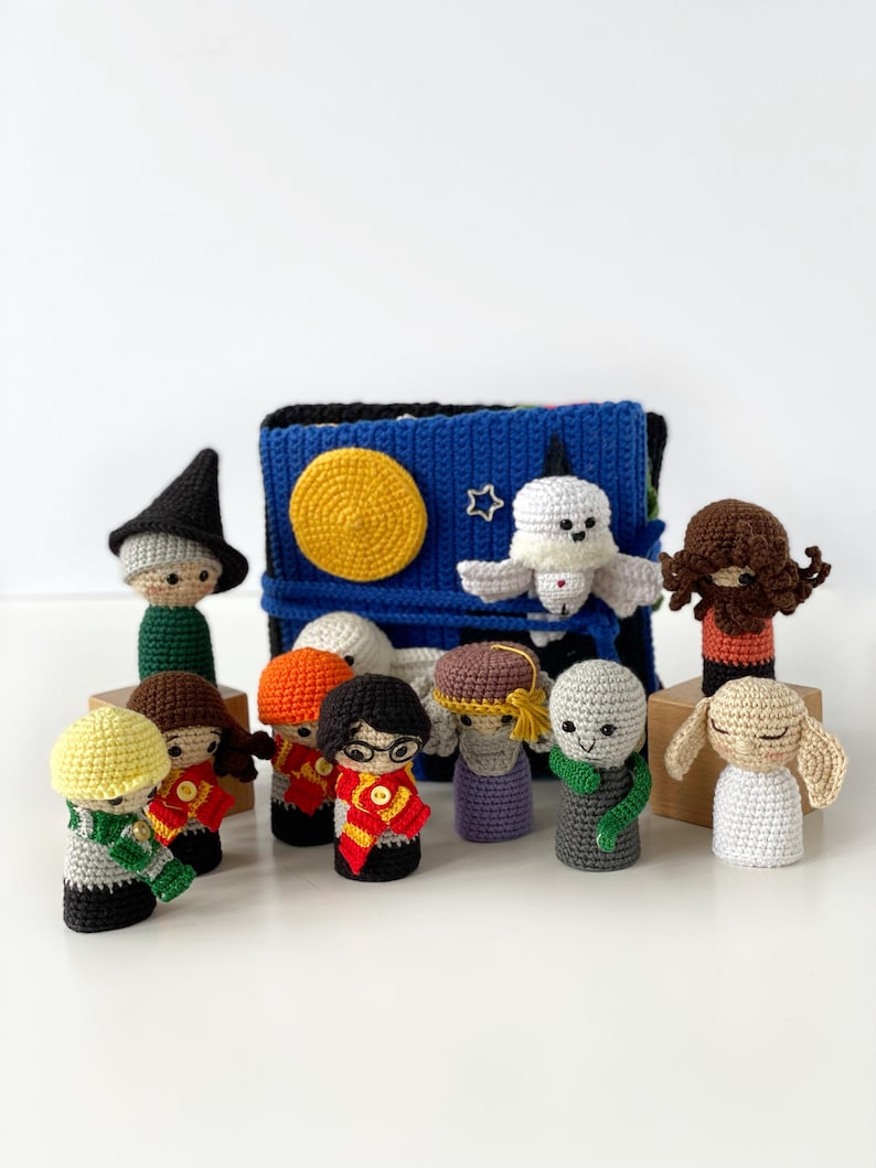 May include: A collection of handmade crochet dolls, each with unique features and outfits. The dolls are arranged in front of a blue and black crocheted book cover with a yellow sun and white star. The dolls are in various colors.