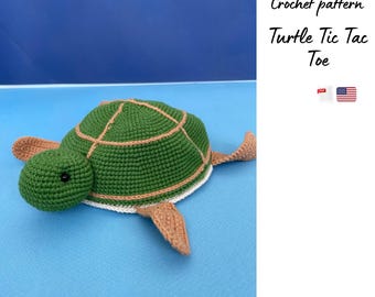 May include: A handmade green crochet turtle with a tan shell pattern and tan flippers. The turtle has a black eye and is on a blue surface. Text on the right says "Crochet pattern Turtle Tic Tac Toe".