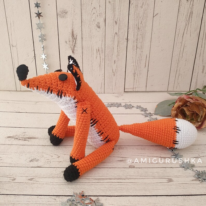 May include: A crocheted orange fox with black eyes and a white belly. The fox has a stitched-on white patch on its back and a white tail tip. The fox is sitting on a wooden surface.