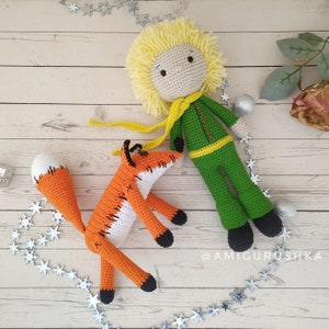 May include: A crocheted fox with an orange and white body and a long tail, lying on a white wooden surface with a crocheted figure of the Little Prince with blonde hair, a green jacket, and a yellow scarf. The Little Prince is standing with his arms at his sides. The background is a white wooden surface with a string of silver stars.