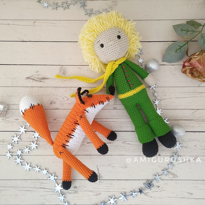 May include: A crocheted fox with an orange and white body and a long tail, lying on a white wooden surface with a crocheted figure of the Little Prince with blonde hair, a green jacket, and a yellow scarf. The Little Prince is standing with his arms at his sides. The background is a white wooden surface with a string of silver stars.