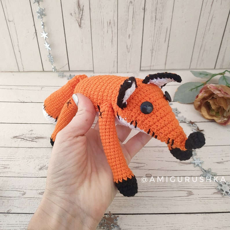 May include: A crocheted orange fox plush toy with black and white details. The fox has black eyes and a black nose. The toy is being held by a hand.