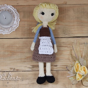 Könnte beinhalten: Eine gehäkelte Puppe mit blonden Haaren, einem braunen Kleid und einer weißen Schürze. Die Puppe trägt schwarze Schuhe und hat einen süßen Gesichtsausdruck. Die Puppe sitzt auf einer Holzoberfläche.