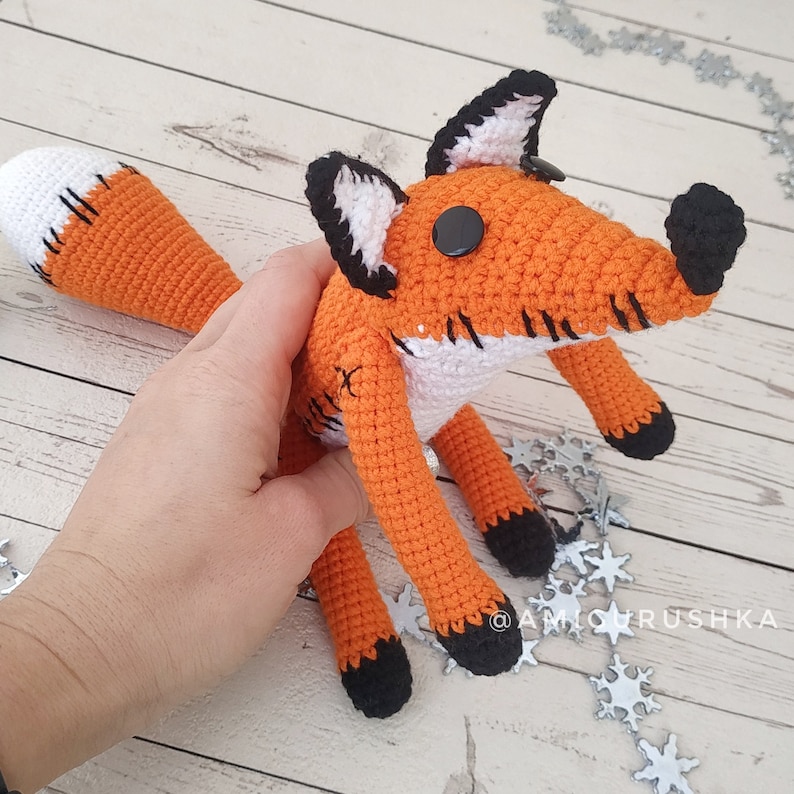 May include: A crocheted orange and white fox plush toy with black eyes and a black nose. The fox is sitting on a white wooden surface with silver snowflake decorations.