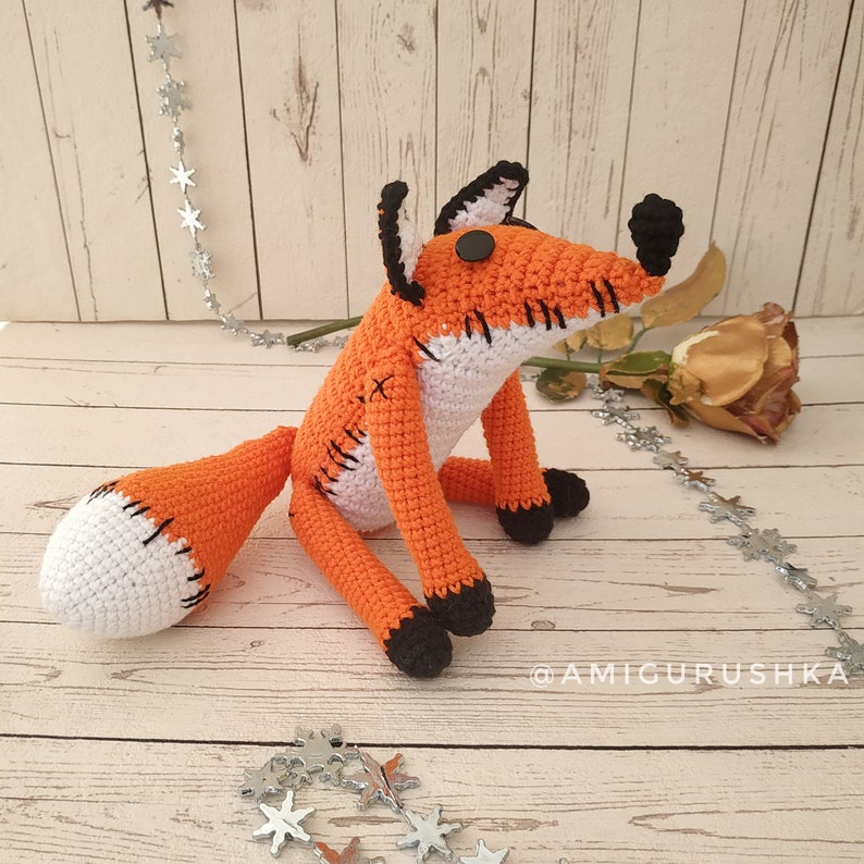 May include: A crocheted orange fox with white accents and black eyes. The fox is sitting on a wooden surface with silver stars scattered around it.