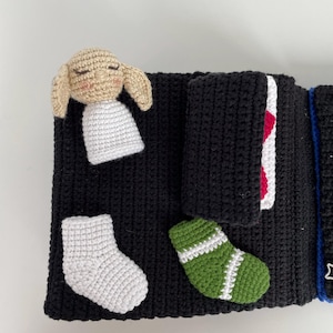 May include: A handmade black crochet book with a beige bunny, white socks, and a green sock with white stripes. The book has a red and white accent and a blue string with a star.