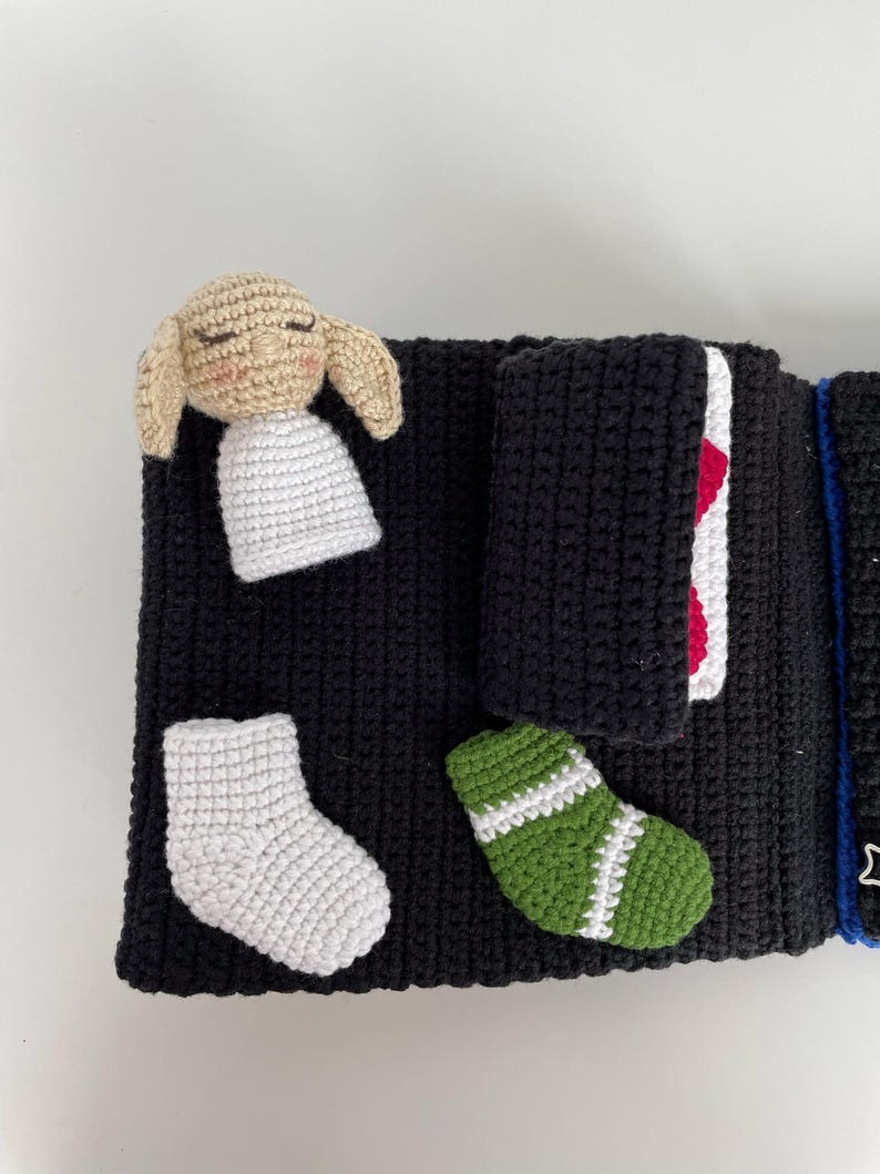 May include: A handmade black crochet book with a beige bunny, white socks, and a green sock with white stripes. The book has a red and white accent and a blue string with a star.