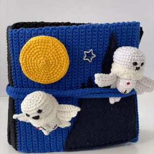 May include: A handmade crochet box with a night sky theme. The box is blue with a yellow moon, a silver star, and two white ghost figures. The box is secured with a blue strap.