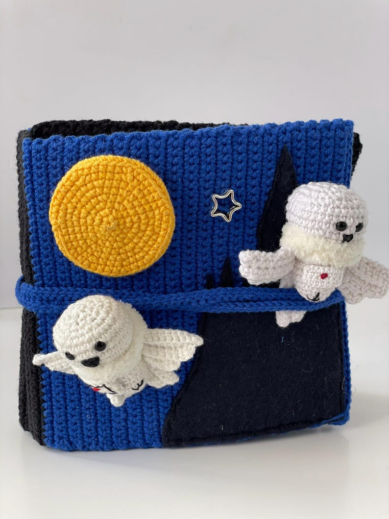 May include: A handmade crochet box with a night sky theme. The box is blue with a yellow moon, a silver star, and two white ghost figures. The box is secured with a blue strap.