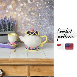 May include: A crocheted teapot and teacup set in white, yellow, pink, and purple. The teapot has a rounded body, a curved handle, and a decorative lid. The teacup sits on a matching coaster. The text "Crochet pattern" is visible.