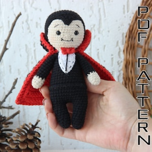 May include: Crocheted amigurumi vampire doll with a red cape, black suit, and white bow tie. The doll has a friendly face with fangs and is holding a small white heart.