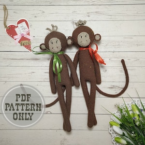 Può includere: Due giocattoli amigurumi di scimmia all'uncinetto con pelliccia marrone, accenti di filato verde e rosso e code lunghe. Le scimmie sono sedute su una superficie di legno bianca.